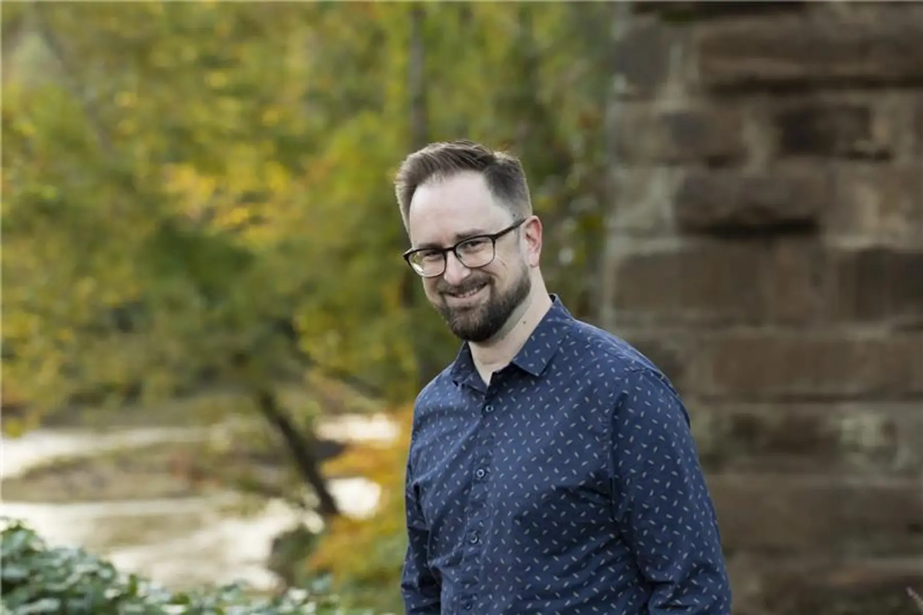 Outdoor portrait of David McCormick, a man with glasses and a beard, smiling in a patterned navy shirt near a stone wall and river.