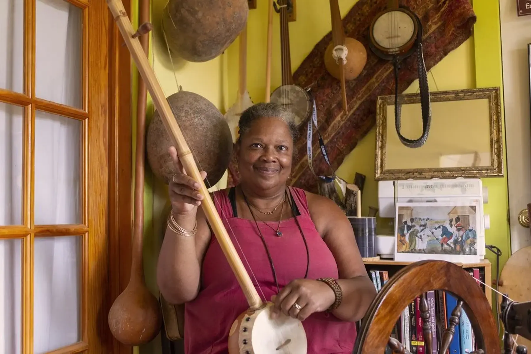 Dena Jennings smiles while holding a gourd banjo, surrounded by a collection of gourd instruments, framed artwork, and books.