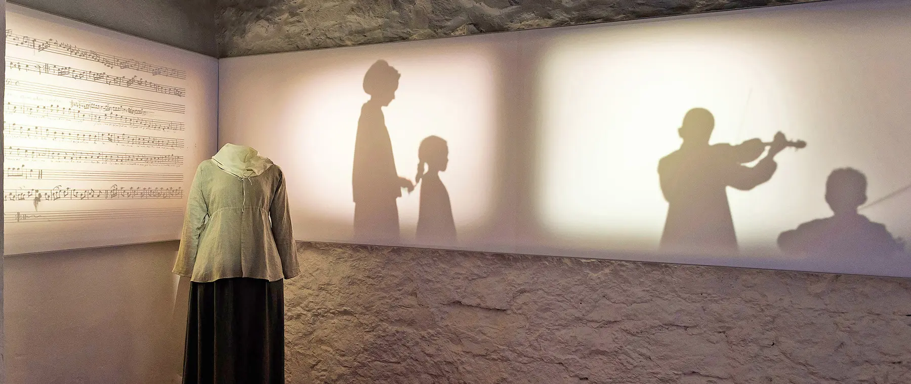 A woman's dress form stands in front of projected shadows depicting Sally Hemings coming her daughter's hair while two boys play the violin#asset:2021@1:alt