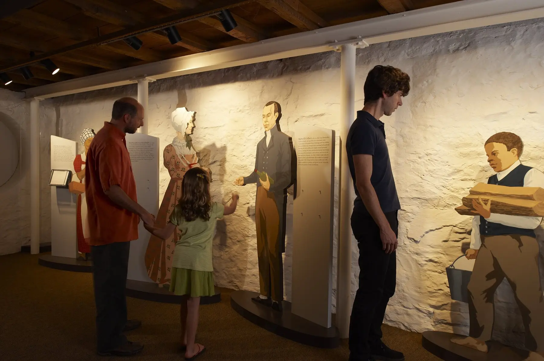 A man and his daughter look at cutout life-sized depictions of a man and a woman in early 19th-century dress while another man looks at a cutout image of an enslaved boy carrying firewood and a bucket.